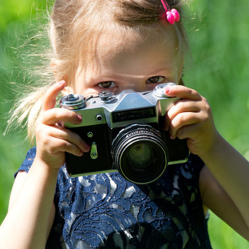 A little girl takes a picture with a retro film camera in the summer in the park. Little pretty child baby girl in light dress holding retro vintage photo camera on green grass in city park.