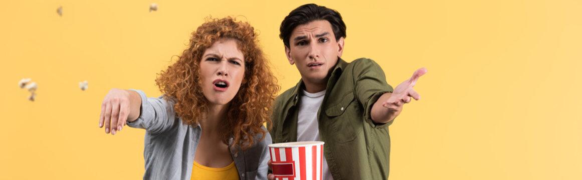 Panoramic Shot Of Emotional Irritated Couple Watching Bad Movie And Throwing Popcorn, Isolated On Yellow