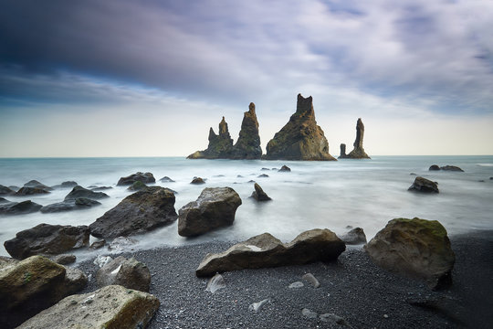 Famous tourist landscape with basalt rock formations Troll toes (Trolls fingers) on black beach. Ocean waves flow around stones. Reynisdrangar, Vik, Iceland, Atlantic Ocean, Europe. Travel postcard.