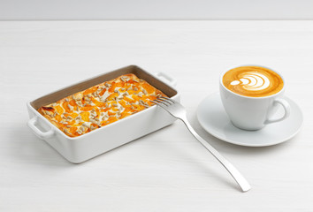 Cup of coffee and homemade cottage cheese casserole with dried apricots and sunflower seeds on white wooden table.