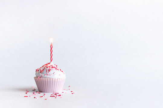 on a white background there is a beautiful cupcake in a pink tin with airy white whipped cream with one hot red and red striped candle i with a beautiful sprinkle of candy