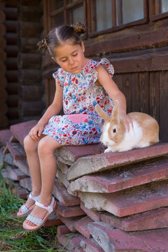 vertical photo of a six year old girl who strokes a rabbit sitting near an old farmhouse