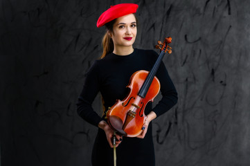 Portrait of a pretty brunette musician girl in a black dress and red beret on a gray background plays the violin