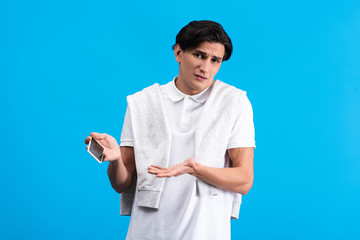 skeptical young man with shrug gesture using smartphone, isolated on blue