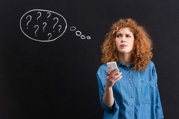 attractive thoughtful woman using smartphone, question marks in thought bubble on blackboard behind