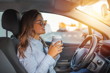 Cheerful woman holding steering wheel and coffe cup while driving a car, close-up view. Woman...