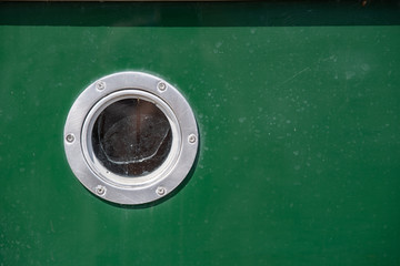 Close-up semi abstract of part of the hull of a green-painted boat with a single round metal and glass porthole window in bright sunlight