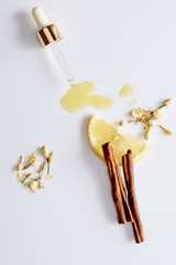 Fototapeta na wymiar Top view of cosmetic oil flowing out of dropper next to slice of lemon, sticks of cinnamon and vanilla buds on white background