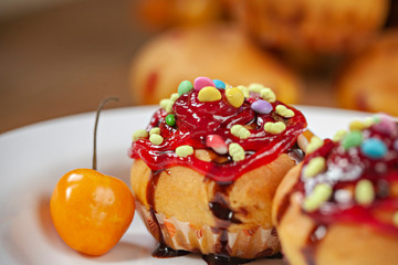 Close-up of garnished muffins with the toppings of sweet candy, jelly and dark chocolate syrup in white plate with poha berries placed on wooden table.