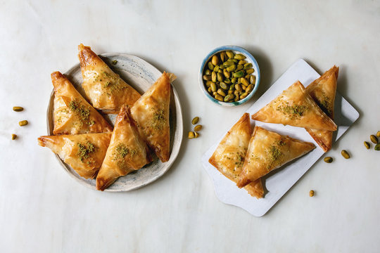 Homemade Turkish traditional dessert baklava with pistachio served on white wooden cutting board with bowl of nuts over white marble background. Flat lay, space