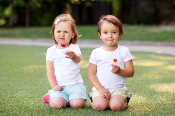 Two little toddler kids, boy and girl, sitting on green grass field or playground (stadium) holding two bright lollipop candies. Summer activities, happy carefree childhood, friendship and fun concept