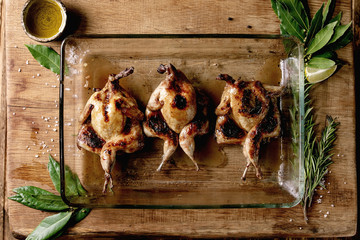 Roasted grilled butterfly quails in glass baking tray with greens, salt and olive oil over wooden background. Flat lay, space