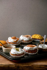 Homemade citrus oranges or clementines sweet muffins cupcakes in baked tray with different cream, pistachio, caramel toppings in bowls above on wooden table. Copy space