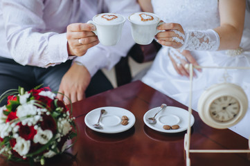 The bride and groom are sitting in a cafe with cups of coffee with foam in the form of a heart and a bouquet of roses and eustomas. Photography, concept.
