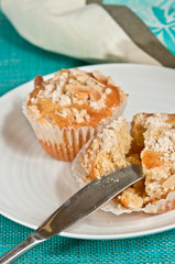 Top view, close up of two freshly baked apricot, almond streusel muffins in paper form, one cut in half with an artisan knife on a round, white plate