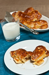 Top view, close distance of a coconut, almond bear claw danish, torn open on a round, white plate with a glass of milk, and an oval dish of four claws,  artisan in background, on a blue place setting