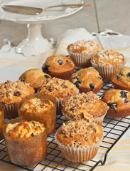 Top view, close distance of three rows of a variety of freshly baked muffins in their fork, cooling on a wire cooling station, with a pedestal dish on a table