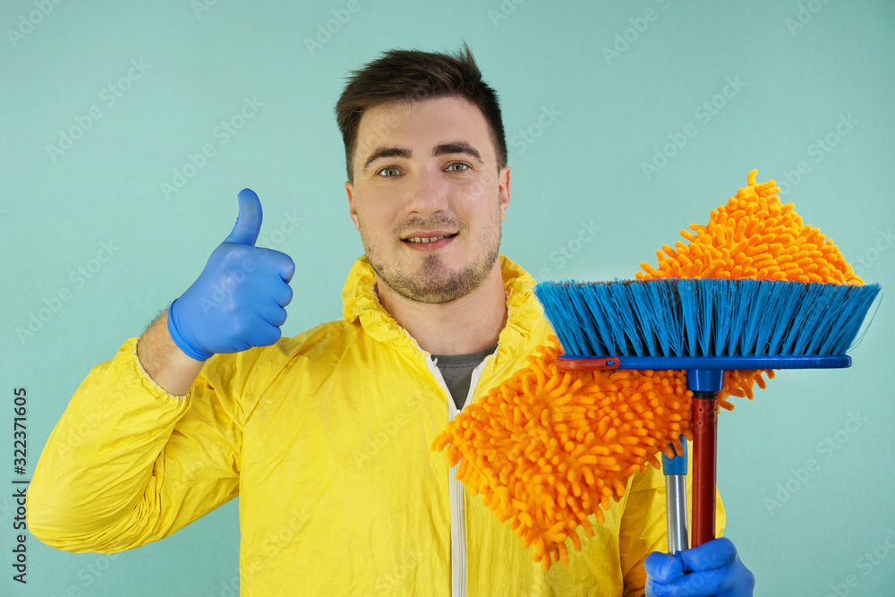 Wall mural cheerful male cleaner with a mop and a broom in his hands in rubber gloves shows a thumbs up gesture