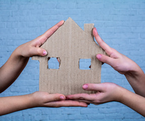 Children's hands hold a house cut out of cardboard. On the background of a white brick wall.