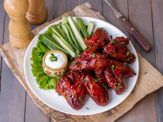 Sticky BBQ Chicken Wings with dipping sauce served hot with cucumber sticks on white plate. Wooden background, selective focus.