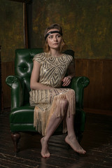 Portrait of young adult beautiful female model in 1920s style golden pattern dress sitting in...
