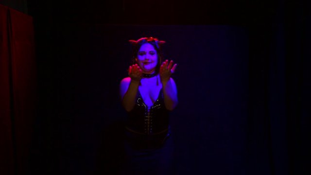 A woman in the form of a devil with an ominous smile dances in red lighting on a black background