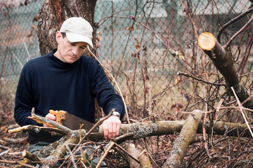 Middle-aged man in spring in the backyard near his house cleans the area from trees that have fallen from the strong wind, the man carves the tree with a hand saw