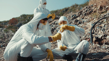 Close-up scientists in chemical suits observing contamined landscape with trash rocks on dump. Low...