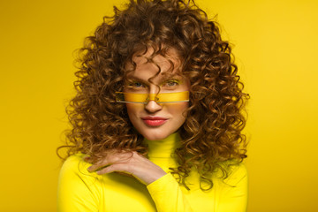 Fashion studio portrait of young beautiful women with curly hair. Nice girl with afro hair and bright make up wearing yellow eyeglasses standing on yellow background. Beauty and hair care concept.