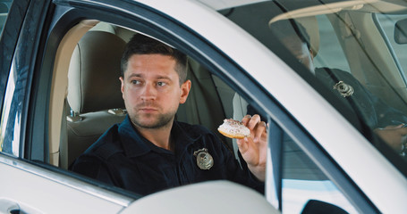 Friendly police officers talking sitting inside the patrol car eating doughnuts together. Two policemen on duty having a tasty break.