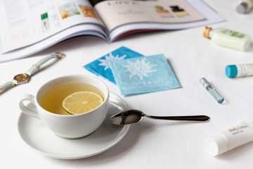 White cup of green tea with lemon on a white bedsheet with aт illustrated magazine, tubes of cream, watch and tea bags. Healthy concept.