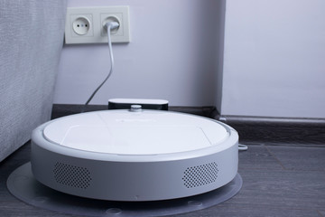 Fototapeta premium white robot vacuum cleaner is charging on the charger on laminate floor cleaning dust in living room interior. Smart electronic housekeeping technology.