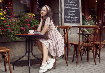 Young beautiful bright girl in a summer dress sitting at a European cafe table
