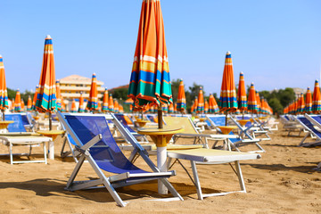 Blue sunbeds with bright multicolored orange green striped umbrellas on beautiful sand beach under blue sky. In front of adriatic sea. Summer holidays, vacation on the coast. Tourism in europe, Italy