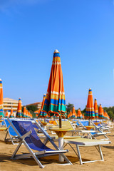 Fototapeta na wymiar Blue sunbeds with bright multicolored orange green striped umbrellas on beautiful sand beach under blue sky. In front of adriatic sea. Summer holidays, vacation on the coast. Tourism in europe, Italy
