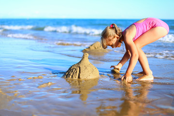 Cute little girl spend holidays in pink one-piece swimsuit, builds sand castle, plays in the water on the shore of the blue sea. Vacation with kids on beach on a warm sunny summer day before sunset