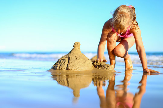 Cute Little Girl Spend Holidays In Pink One-piece Swimsuit, Builds Sand Castle, Plays In The Water On The Shore Of The Blue Sea. Vacation With Kids On Beach On A Warm Sunny Summer Day Before Sunset