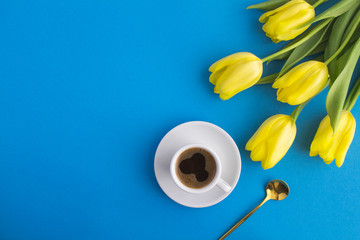 Black coffee in the white cup and yellow tulips on the blue background. Top view. Copy space.