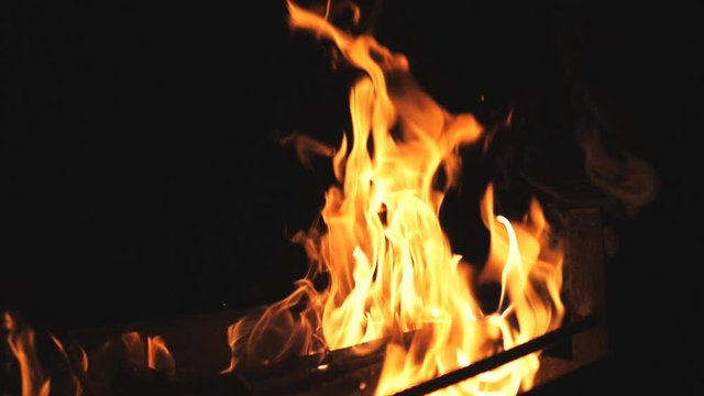 Close up of beautiful flames rises from burning slices wood in mangal at night. Bonfire inside metal brazier at darkness. Firewood burns in barbecue on black background. Concept of warmth and rest