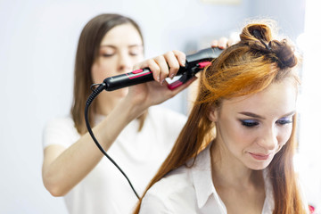 Hairdresser in white t-shirt is spraying hair of young redhead model with bright make-up with liquid from a bottle, making a hairstyle. Close up