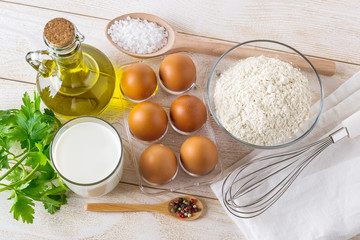 Ingredients prepared for cooking classic omelette: brown eggs, milk, flour, olive oil, salt, pepper and parsley on a white wooden table. Easy preparation of breakfast.