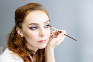 Obraz premium Redheaded female in white shirt and jeans is holding a make-up palette, applying make-up with brush, sitting in front of mirror. Close up