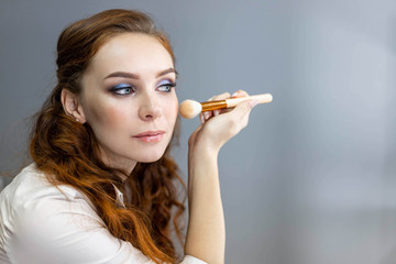 Obraz premium Redheaded female in white shirt and jeans is holding a make-up palette, applying make-up with brush, sitting in front of mirror. Close up