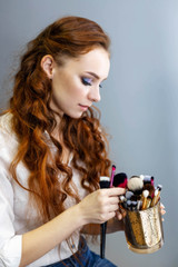 Smiling, ginger girl in white shirt and jeans is choosing brushes while posing sideways sitting...