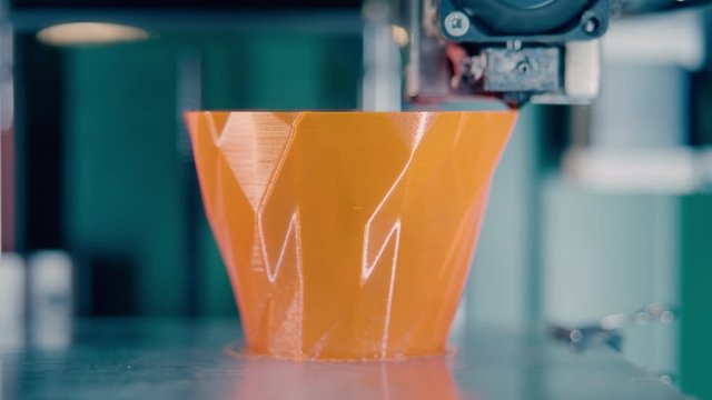 3D-printing machine is producing an orange vase. Three dimensional 3d printer working at 3d printing lab.