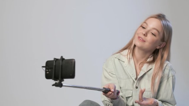 cute female records video by smartphone attached to selfie stick. blond girl filming vlog speaking, smiling, chatting with followers, using cellphone on white background. Beauty blogger recording blog