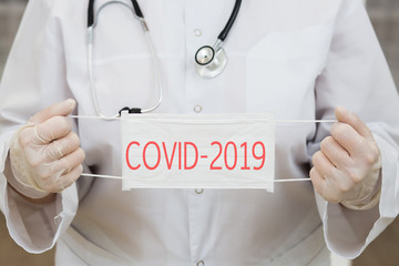 a woman doctor in a white coat, with a phonendoscope around her neck, gloves, and a medical mask in her hands. the inscription on the mask warns of the covid-2019 virus epedemia