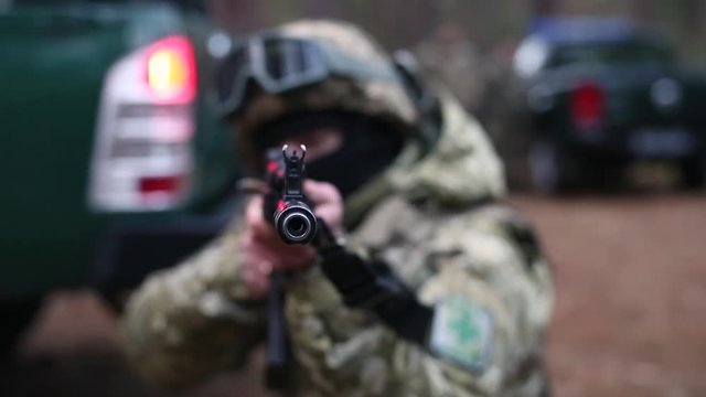 Soldier is tracking a target. Soldier aims from a machine gun directly at the camera. Against a blurred background with cars and soldiers. Armed military man soldier shooting a machine gun.