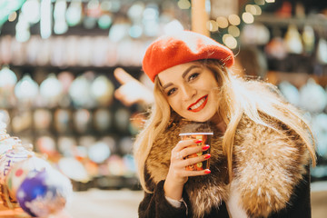 A smiling young woman drinks a warm drink, coffee, tea or toast at the Christmas market. In the background, lights flicker.