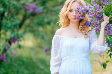 Portrait of wonderful girl with fair hair in long blue dress smells lilac flowers